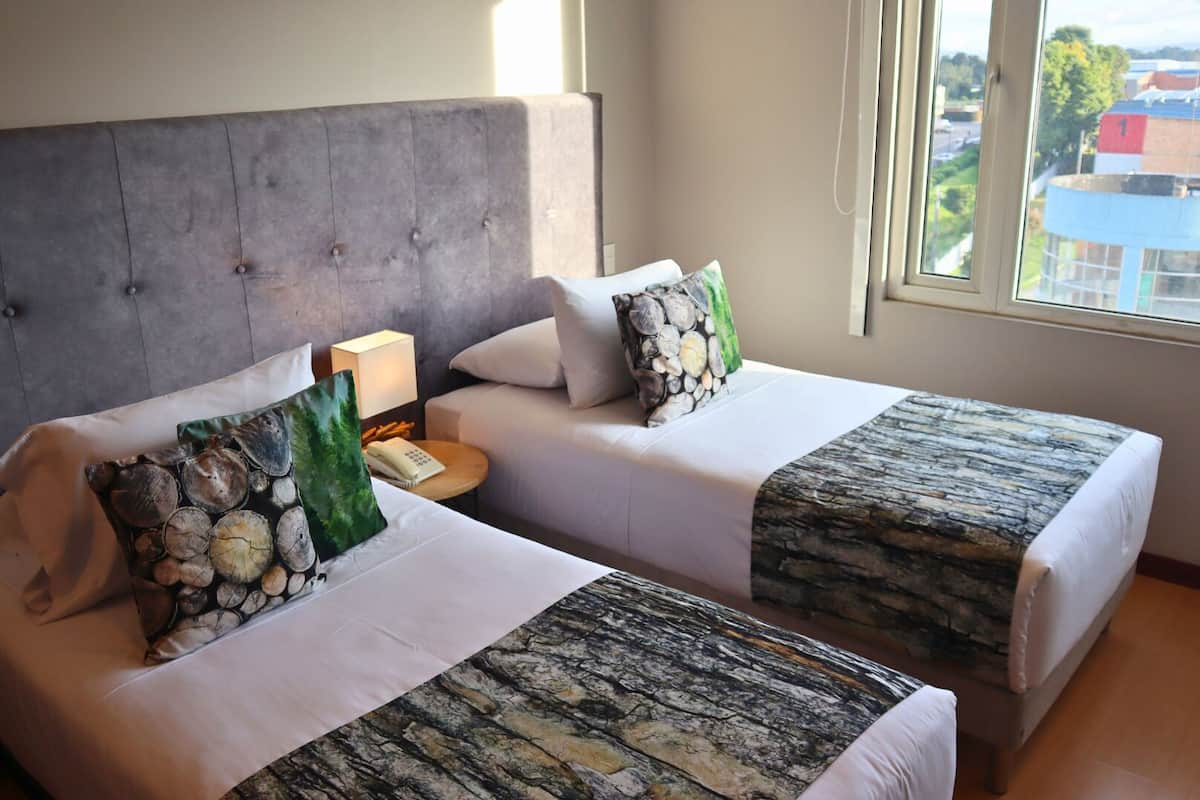Une chambre d’hôtel avec deux lits, chacun doté d’une tête de lit grise et de linge de lit blanc. On y trouve une petite table de chevet avec une lampe et un téléphone. La chambre dispose d’une grande fenêtre offrant une vue sur les bâtiments et les arbres.
