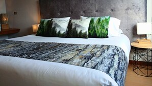 Una cama bien hecha con un cabecero gris, dos almohadas con estampado de bosque y una colcha estampada.