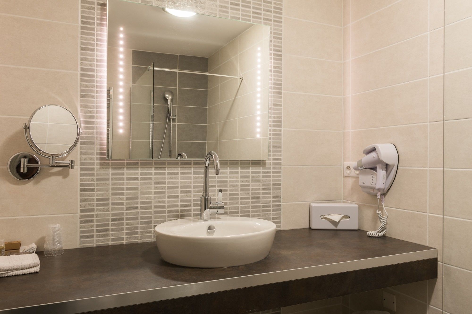 A bathroom with a round mirror, a sink, a handheld showerhead, and a hairdryer mounted on the wall.