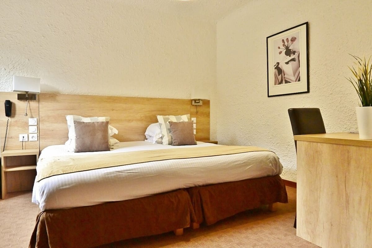 A hotel room with a wooden headboard, a bed with white bedding, a desk with a chair, a bedside lamp, and a framed picture on the wall.