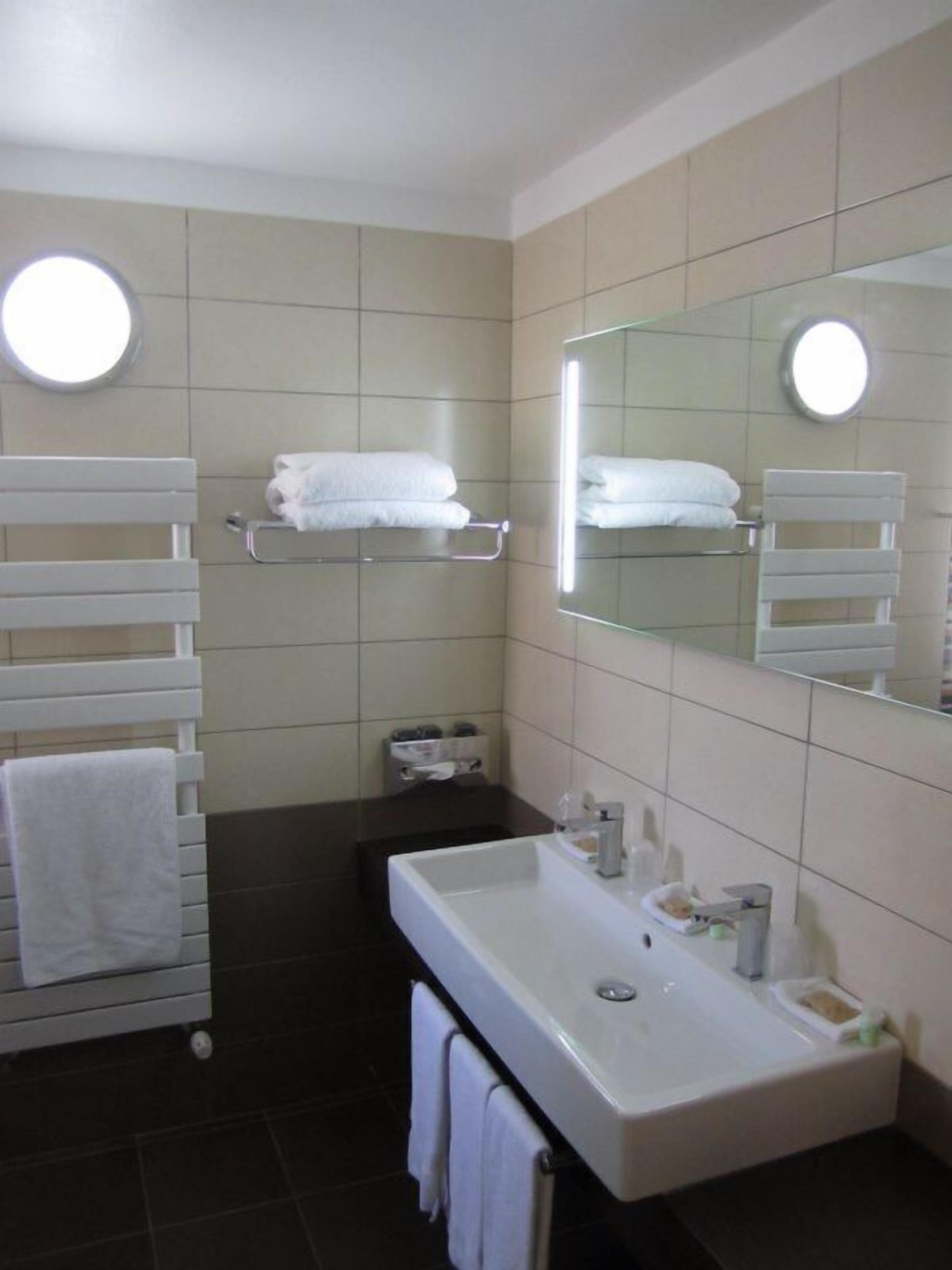 Un baño moderno con un lavamanos blanco, encimera oscura y dos toalleros con toallas blancas.