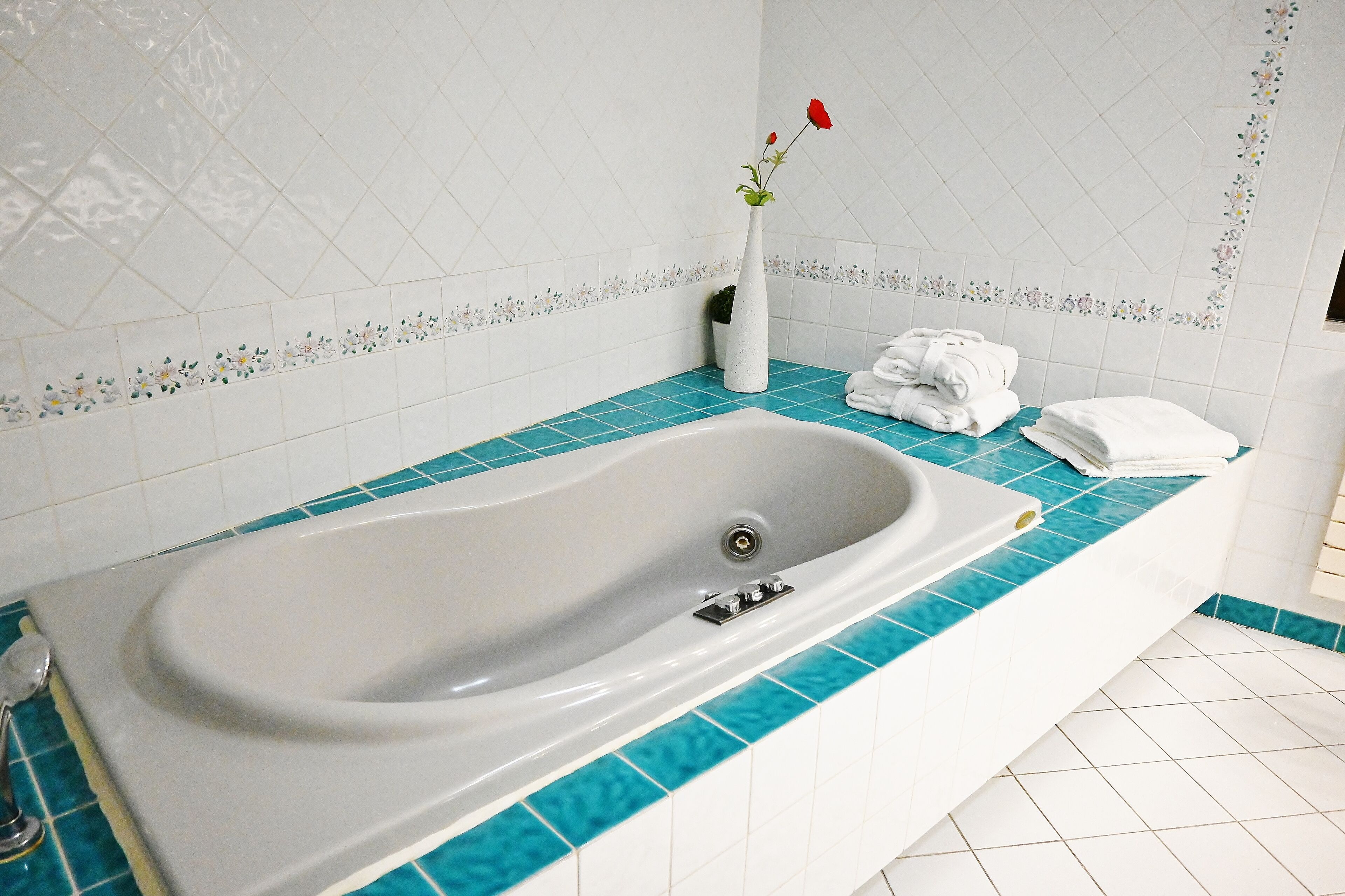 A bathroom with a white bathtub, blue-tiled countertop, white tiles with floral borders, a white vase with a red flower, and neatly folded white towels.