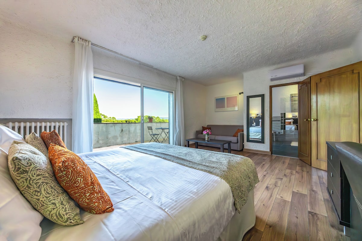 A bedroom with a large bed, a sofa, a dining table, and a view of the outdoors.
