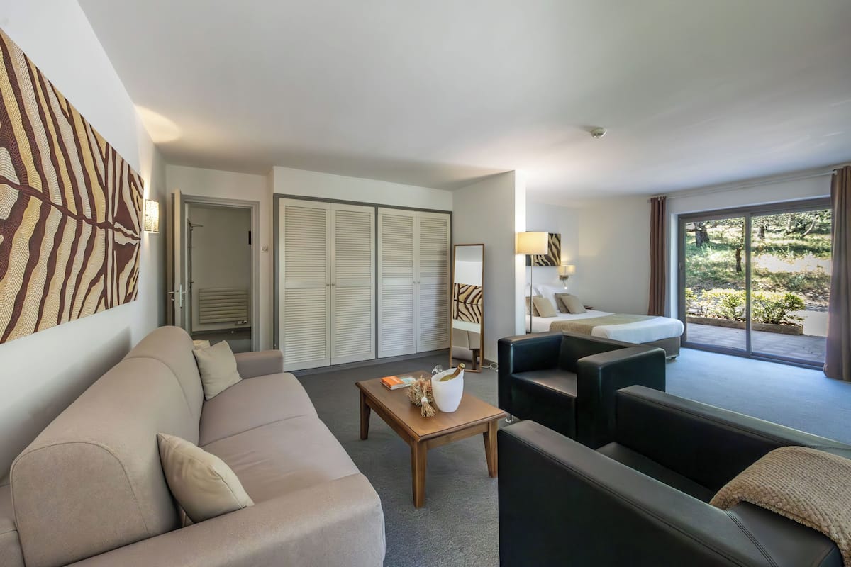 A modern hotel room with a sofa, a coffee table, a bed, and a view of greenery outside.