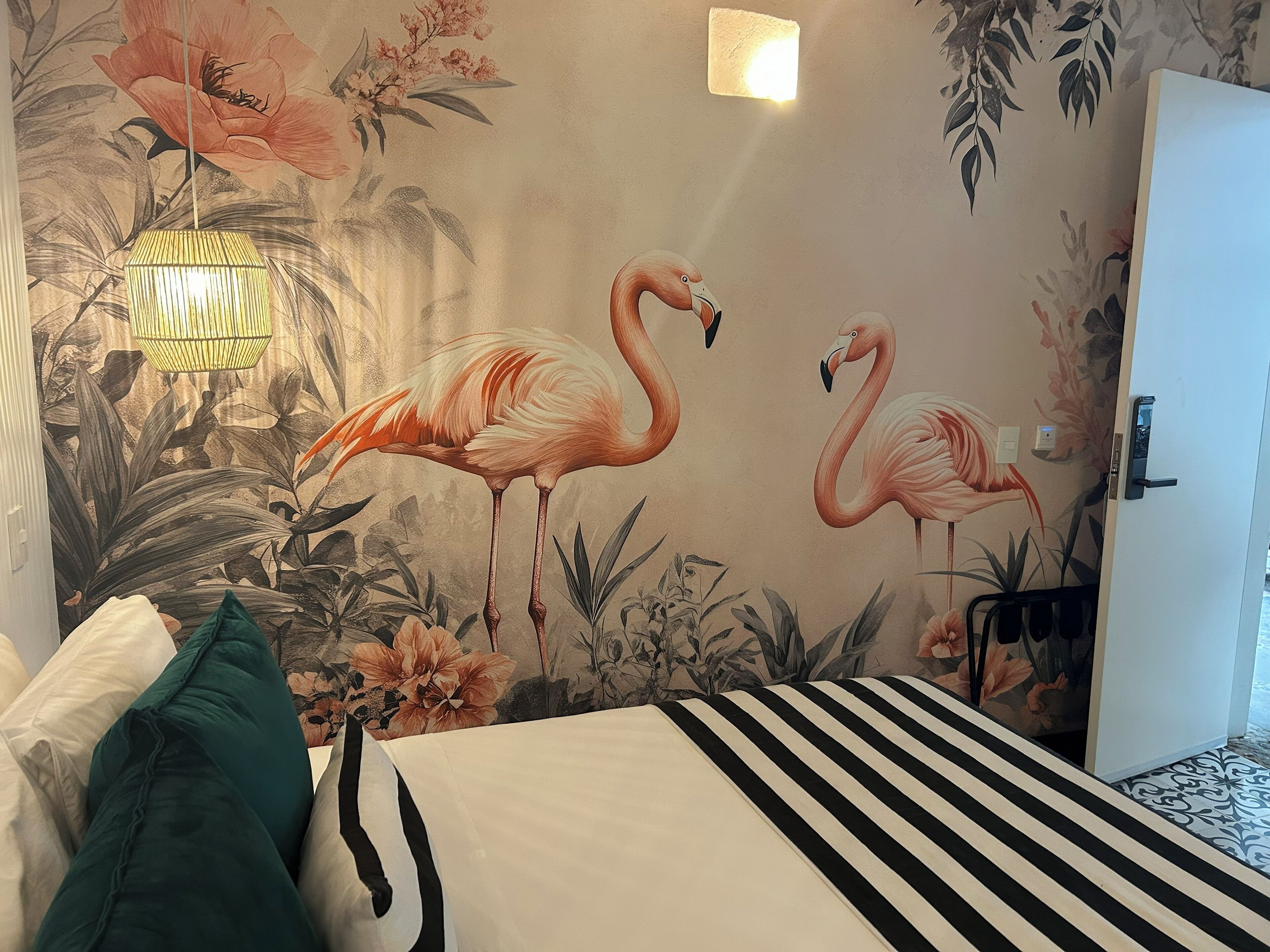 Huone, jossa on sänky, tyynyjä, seinämaalaus, jossa on kuvattuna flamingoja, riippuvalaisin ja lukollinen ovi.