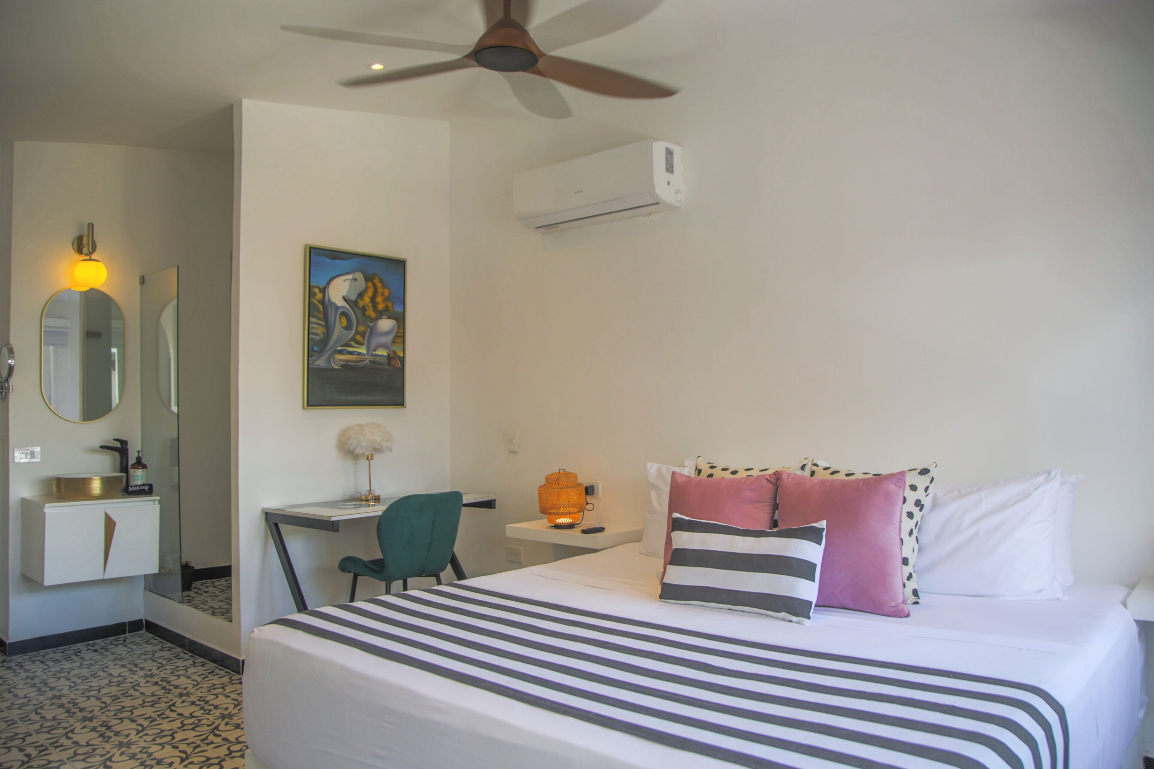 A hotel room with a bed, a desk, a chair, a mirror, a sink, and an air conditioning unit.