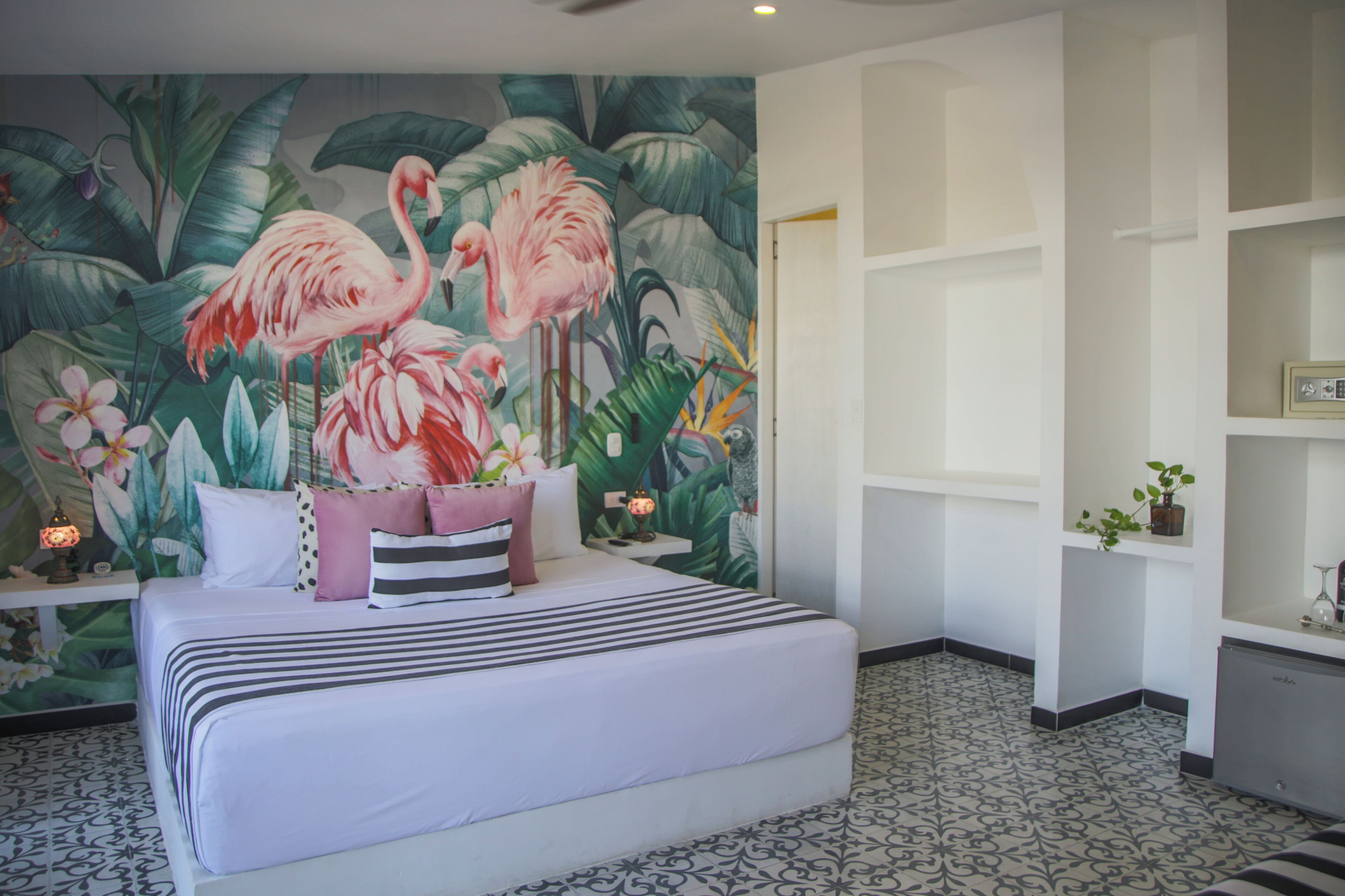 Un dormitorio con cama, mesita de noche, un mural en la pared y estante con una planta.