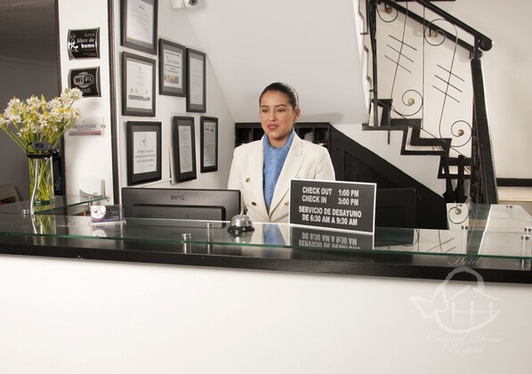 A reception desk with a sign indicating check-in and check-out times, framed certificates on the wall, and a vase with flowers. - Central House Hotel (Bogotá)