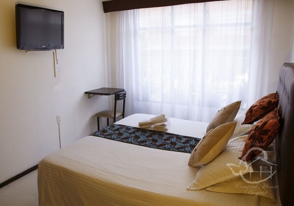 A hotel room with a bed, a TV mounted on the wall, a small table, and a window with sheer curtains.