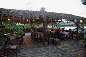 An outdoor dining area with hexagonal stone tiles, wooden tables and chairs, and a thatched roof structure.