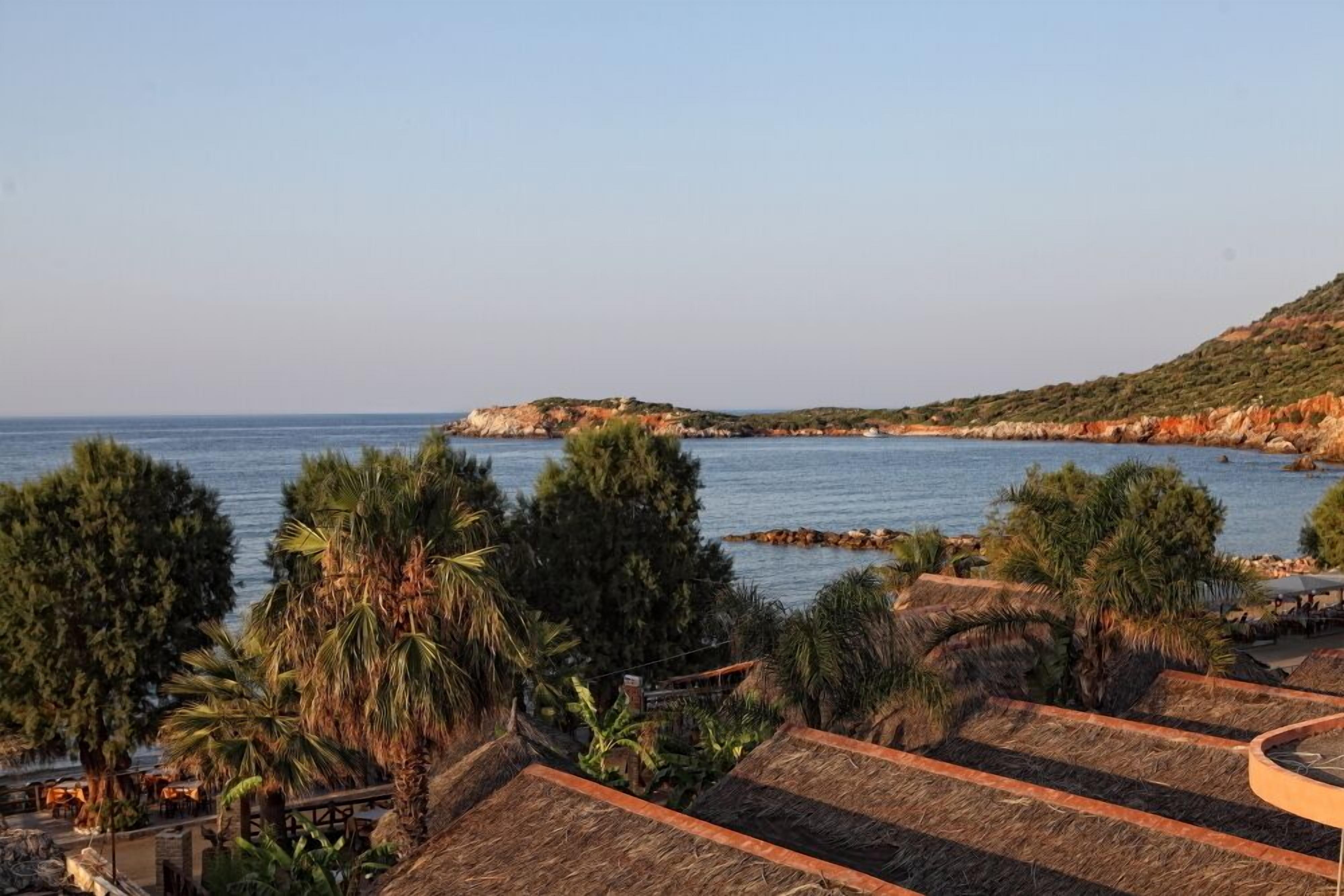 Vista costera con edificaciones de techo de paja, una playa y una isla rocosa en la distancia.