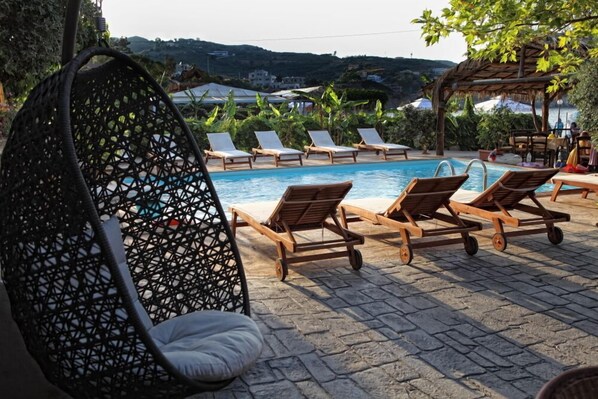 A poolside area with lounge chairs and a hanging chair, surrounded by greenery and a view of hills.