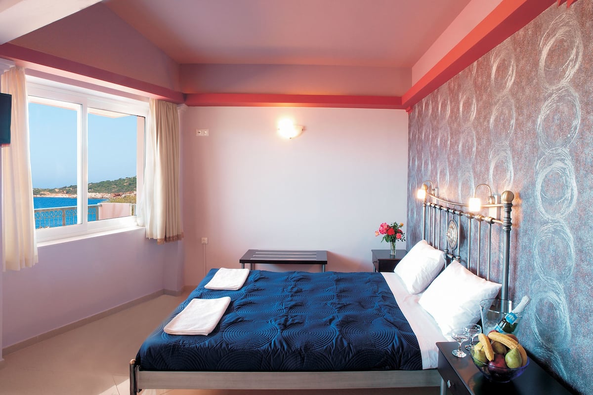 A bedroom with a bed, a TV, a nightstand, a vase with flowers, and a view of the sea.