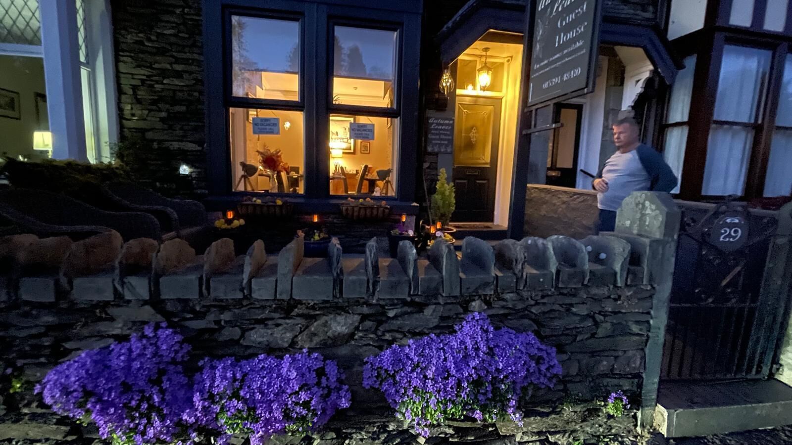 Un muro de piedra con una hilera de estatuas de ovejas y flores moradas frente a un edificio con un letrero que dice "The Guest House".