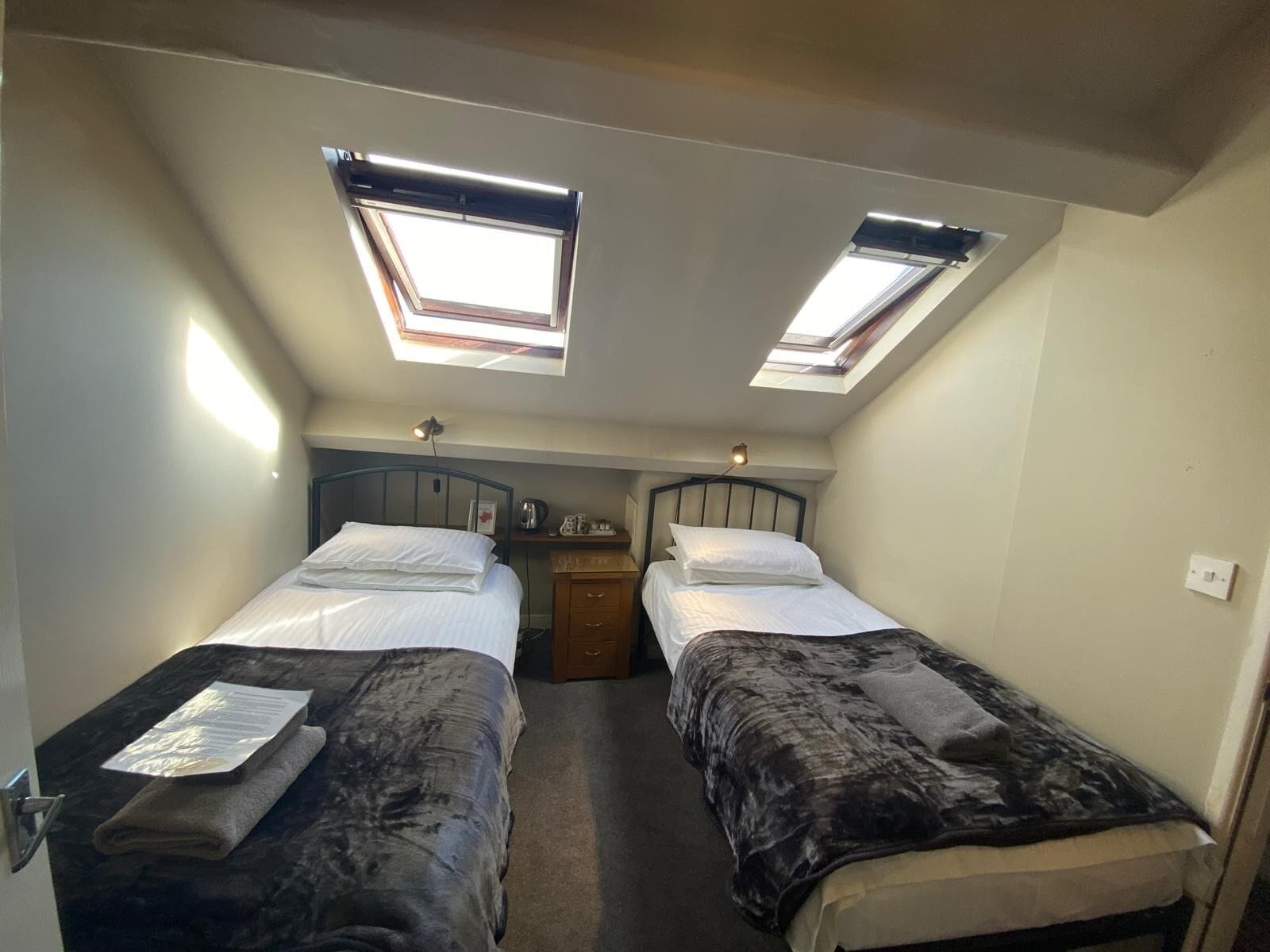 A compact room with two beds, each with a headboard and a bedside table. There are skylights providing natural light.