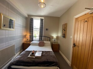 A bedroom with a bed, two bedside tables with lamps, a window with curtains, a radiator, a wooden door, and a painting on the wall. - Autumn Leaves Guest House (Windermere)