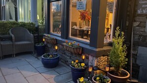 Exterior de un hotel con mesas al aire libre, plantas en macetas y un cartel que indica habitaciones disponibles.