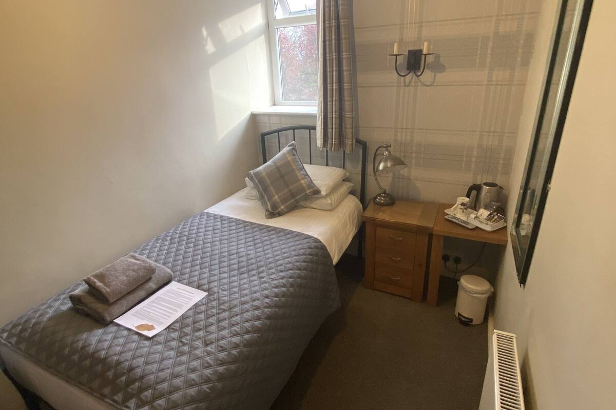 A small, neatly arranged bedroom with a single bed, a bedside table, a lamp, and a window with curtains.