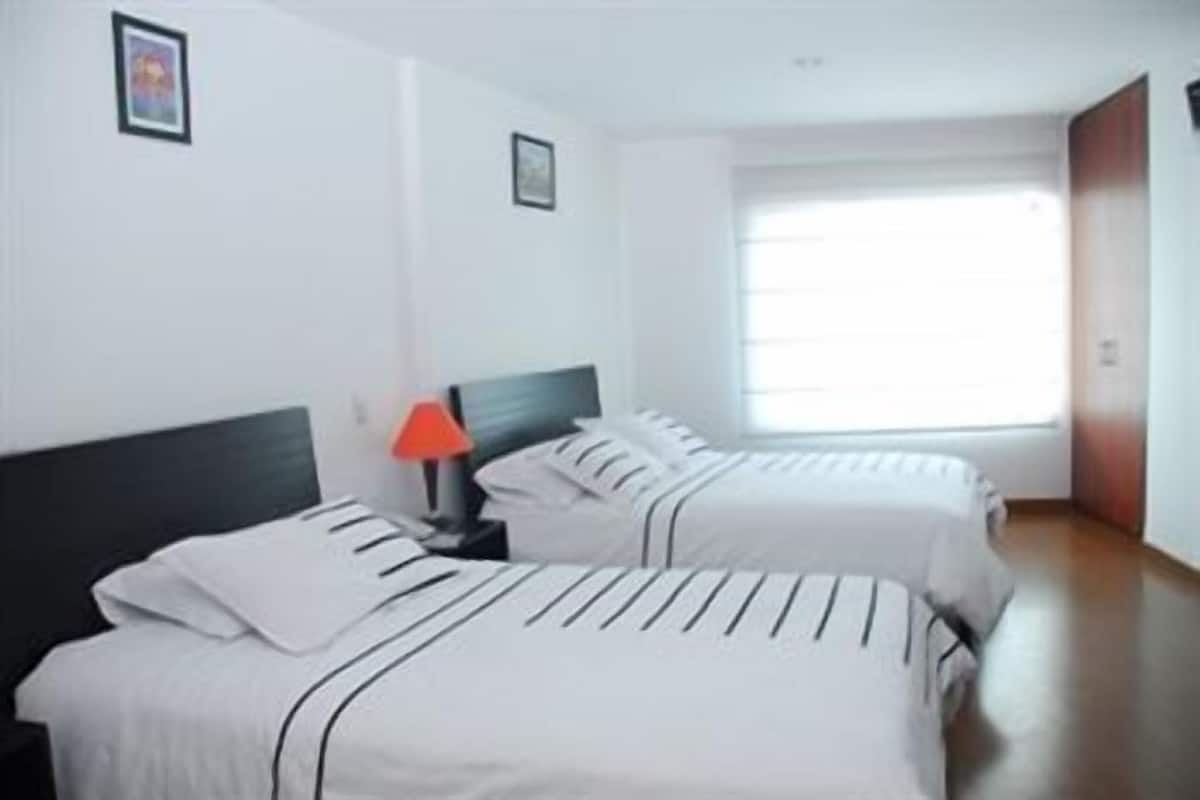 A hotel room with two beds, a nightstand with a lamp, and a window with blinds.