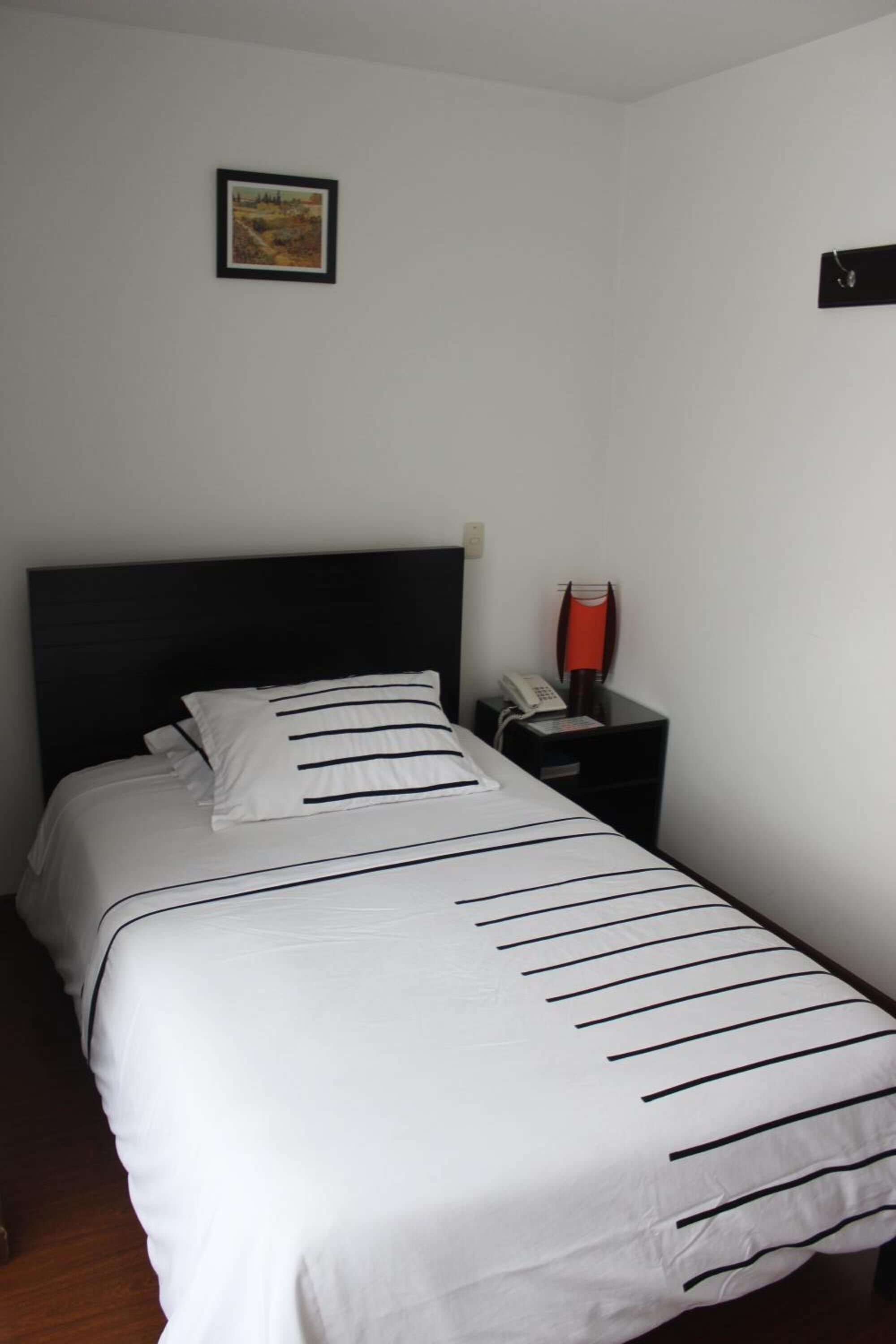 Uma cama bem arrumada com colcha listrada, cabeceira preta e uma pequena mesa de cabeceira com telefone e abajur vermelho.
