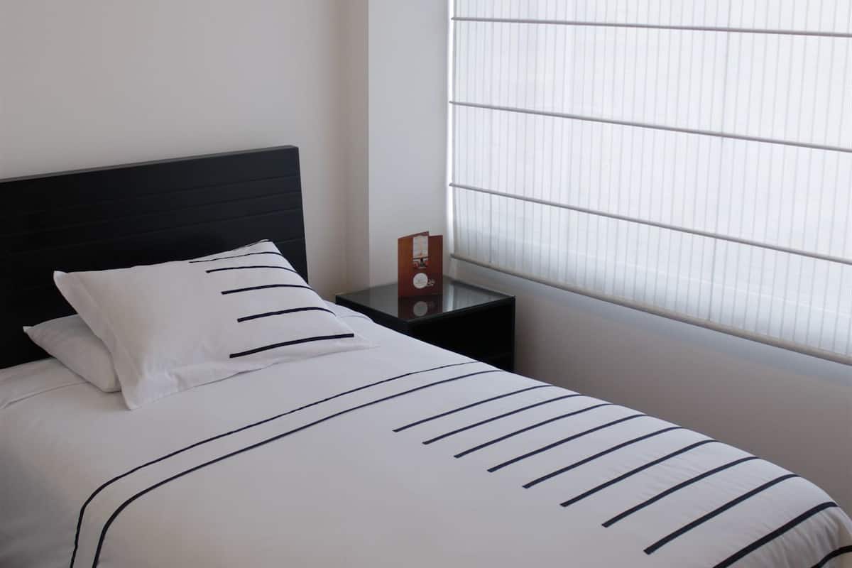 A neatly made bed with white bedding and dark stripes, a black headboard, a bedside table with a clock, and a window with blinds.