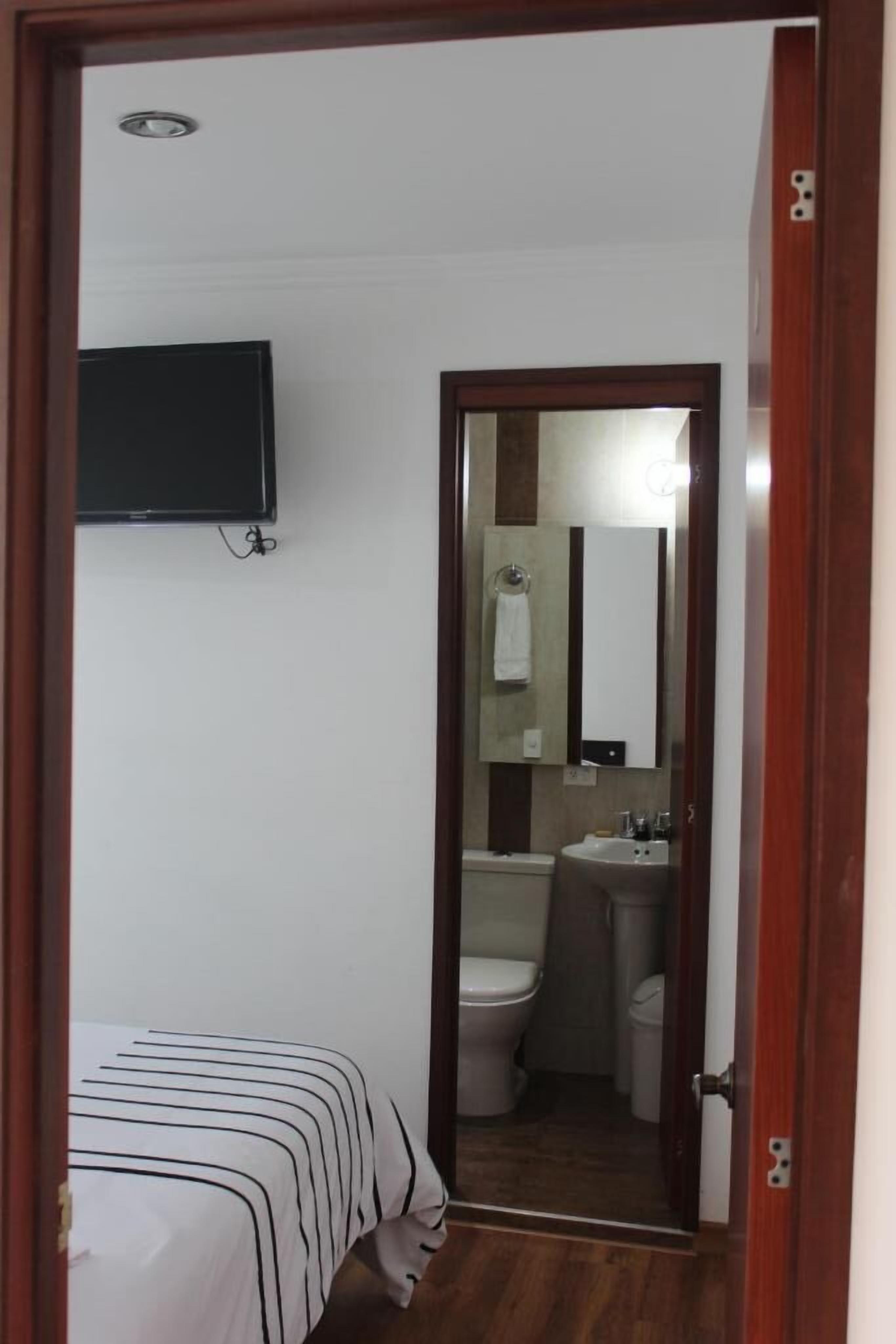 Una camera da letto con un letto, una televisione fissata al muro e una porta in legno che conduce al bagno, dove si trova il water, un lavandino e uno specchio.