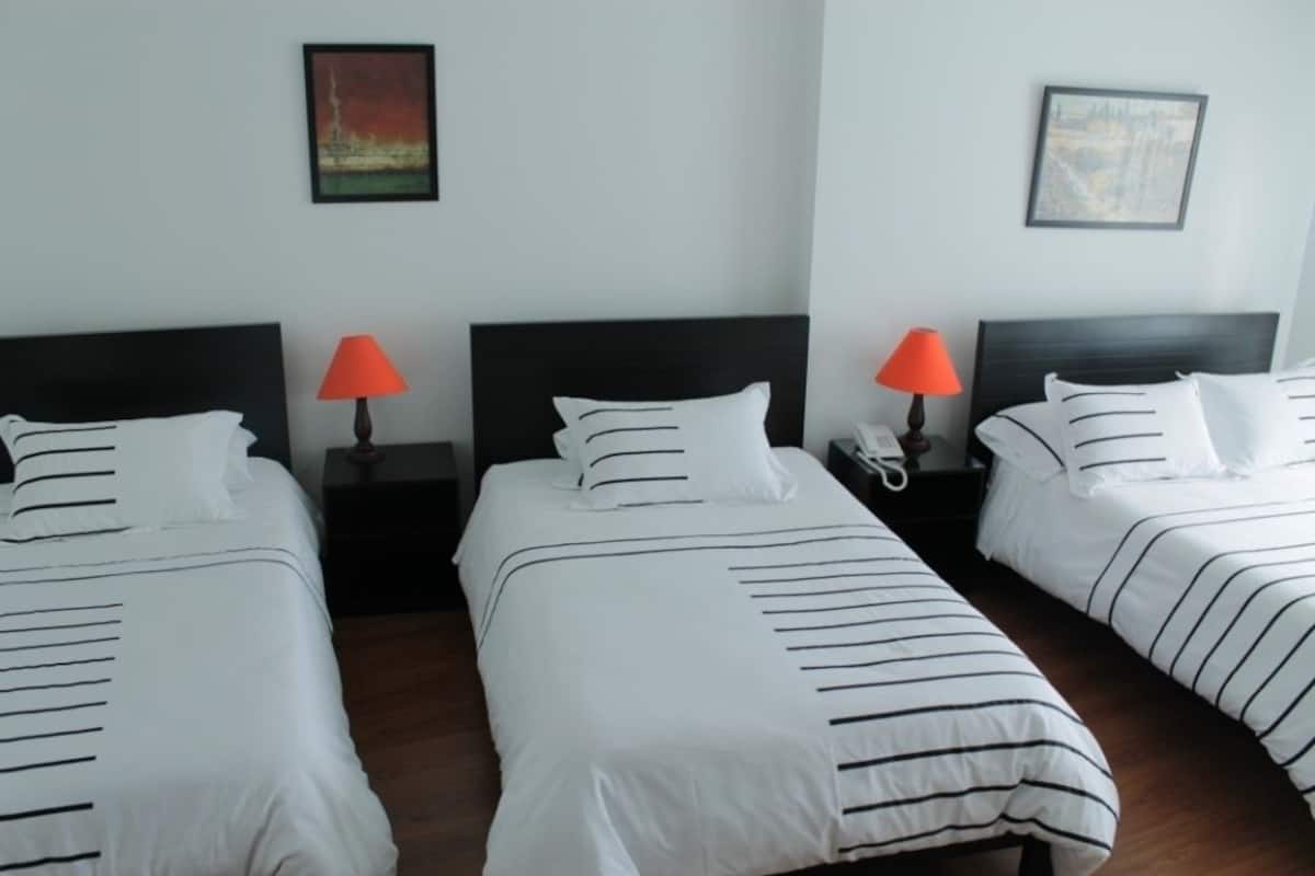 A hotel room with three beds, each with striped bedding and a black headboard. There are two bedside tables with lamps, and artwork is visible on the walls.