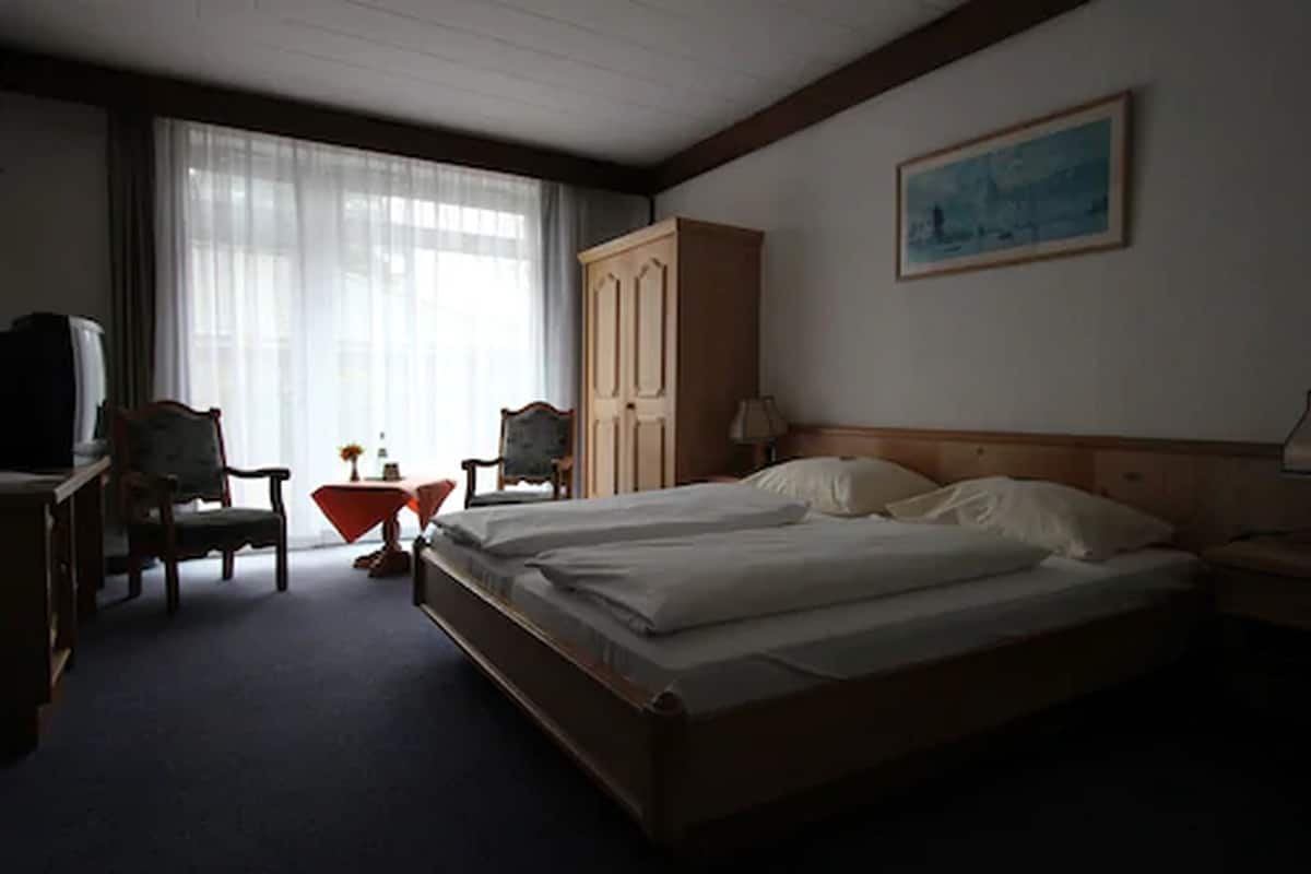 A hotel room with two single beds, a wooden wardrobe, a desk with a chair, a television, and a window with sheer curtains.