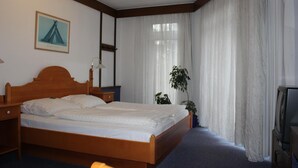 Een slaapkamer met een houten bed, een televisie, een nachtkastje, een lamp, een raam met dunne gordijnen en een ingelijst schilderij aan de muur.