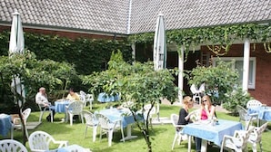 Een terras buiten met tafels en stoelen, omgeven door groen en een bakstenen gebouw.