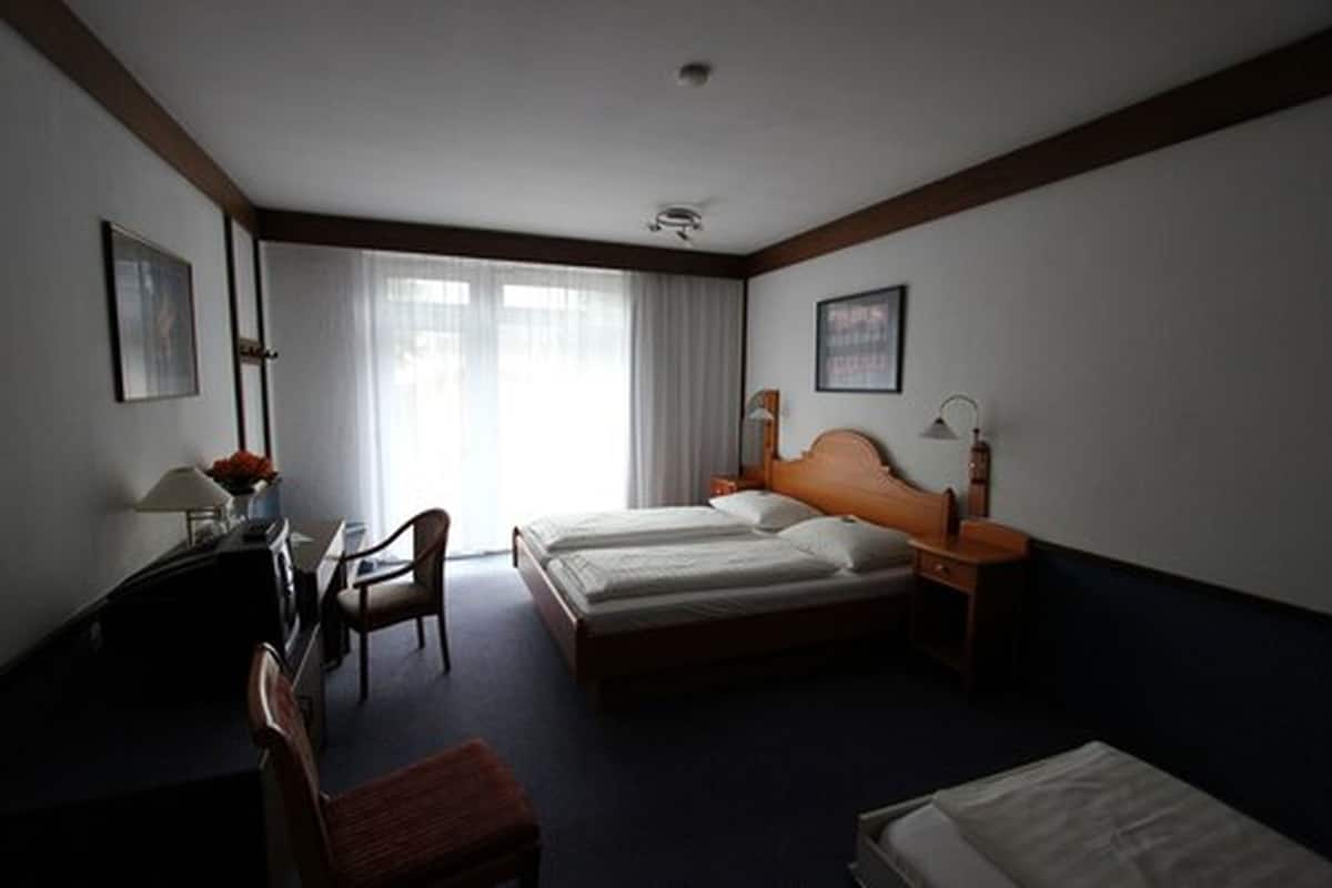 A hotel room with a double bed, a desk with a chair, a small table with a lamp, and a window with curtains.