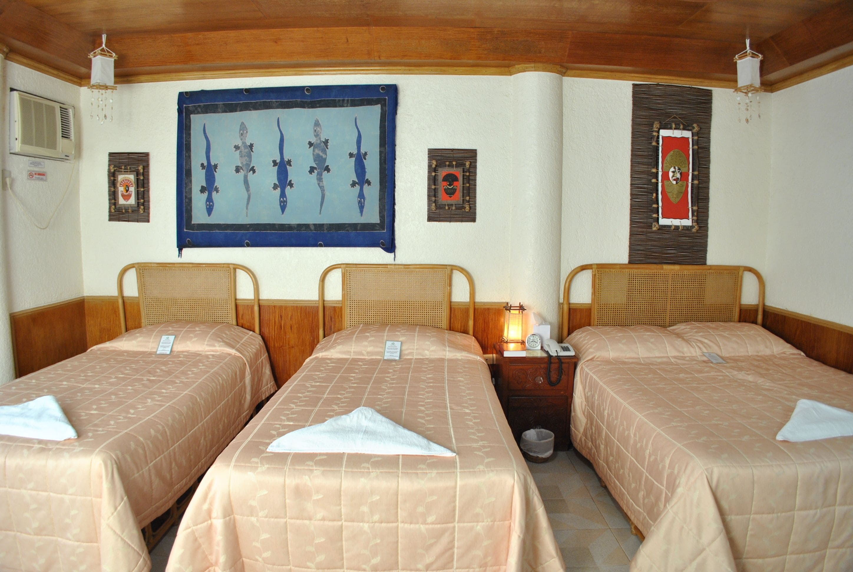 A hotel room with three single beds, each with a beige bedspread and a white pillow. There are wall-mounted air conditioning units on the left wall, and a framed artwork above the beds.