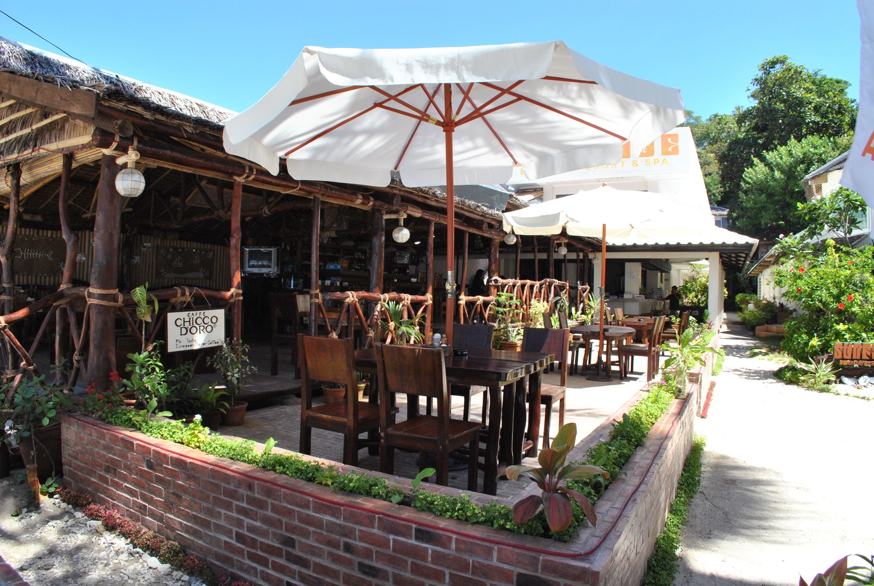 An outdoor seating area with wooden tables and chairs, surrounded by potted plants and a brick wall.