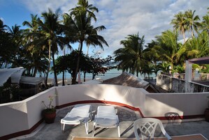 A balcony with two white lounge chairs, a potted plant, and a view of the beach and palm trees.