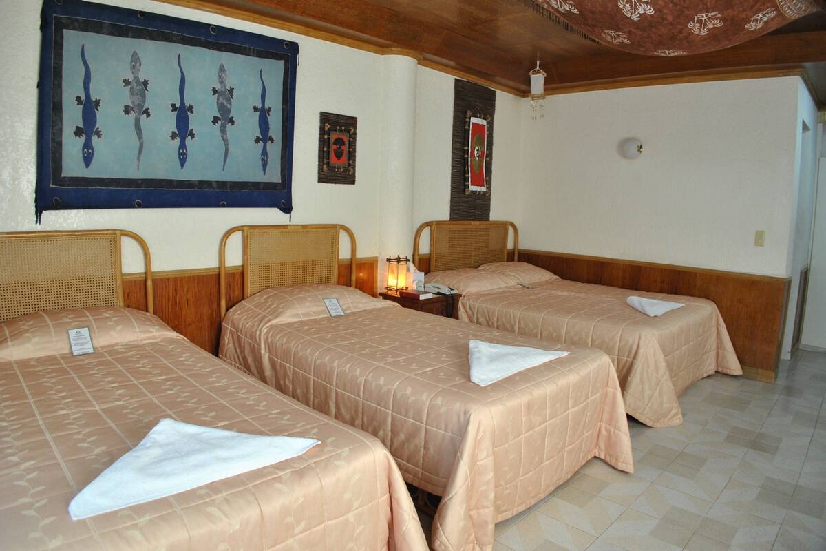 A hotel room with three single beds, each with a beige bedspread and a white pillow. There is a wall-mounted artwork depicting lizards, a framed picture, and a small table with a lamp.