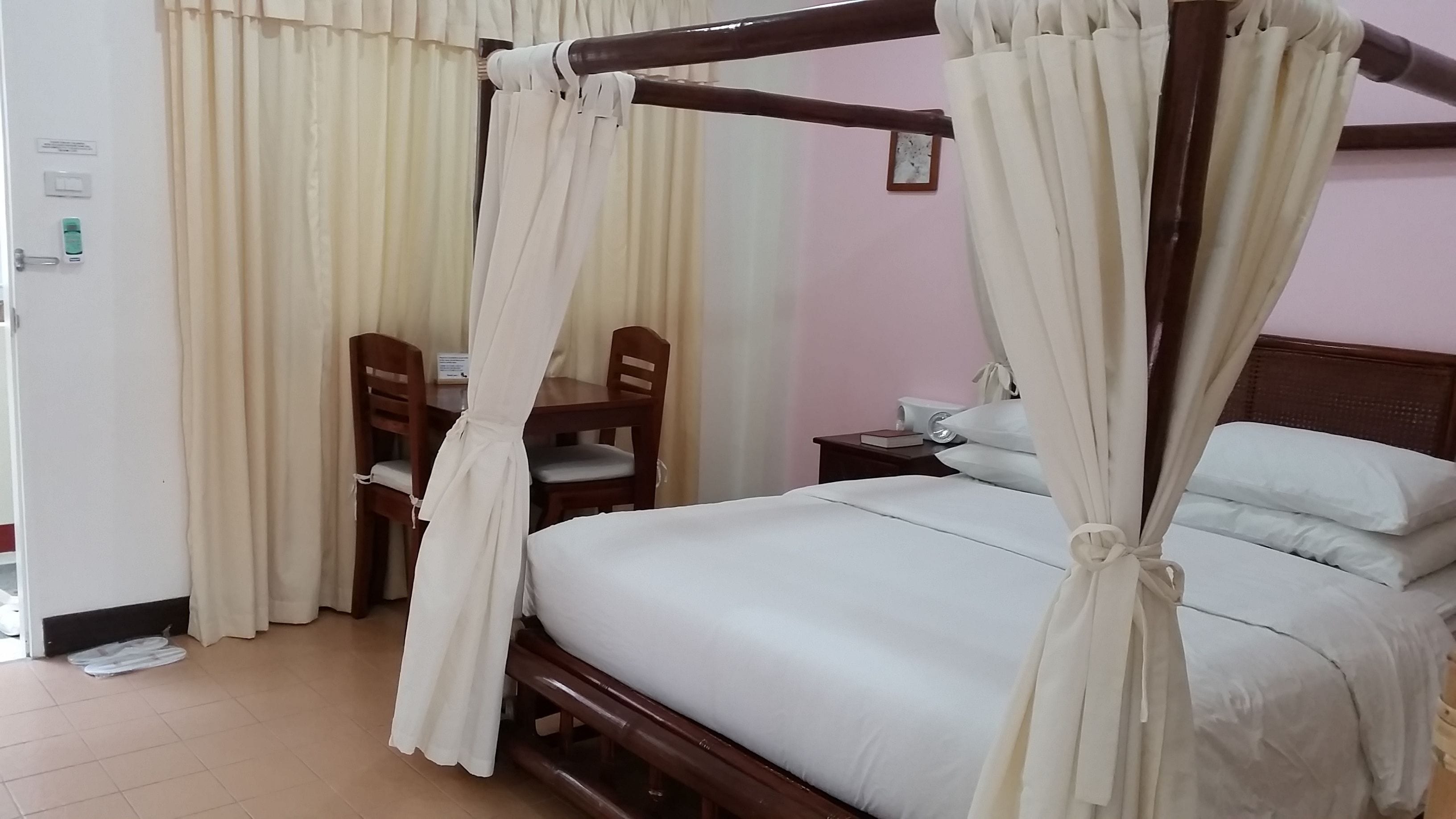 A four-poster bed with white linens, a wooden nightstand with a lamp, and a chair with a cushion.