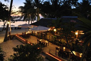 Un restaurante frente a la playa con terraza al aire libre, palmeras y vistas al océano.