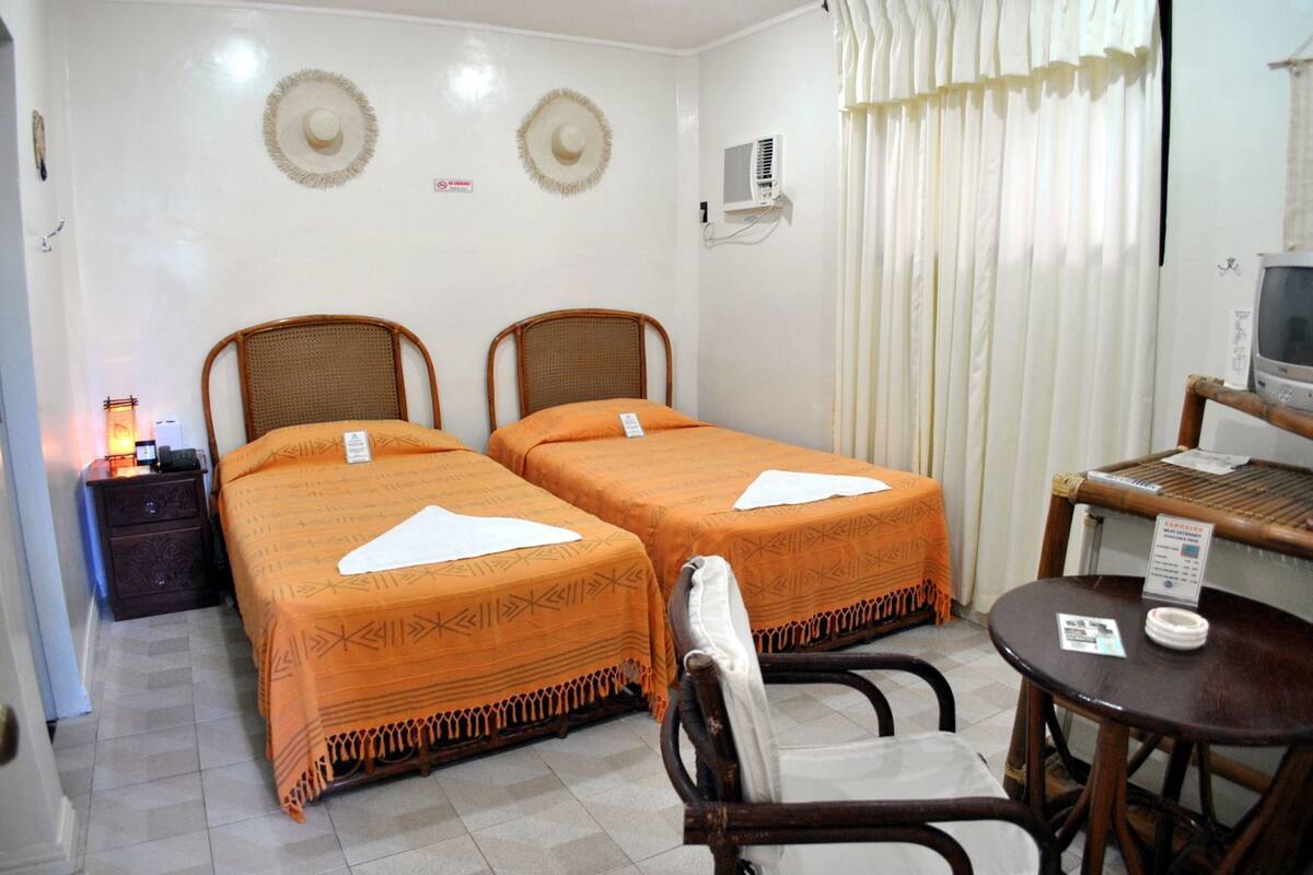 A hotel room with two beds, a small table, a chair, a television, and a wall-mounted air conditioner.