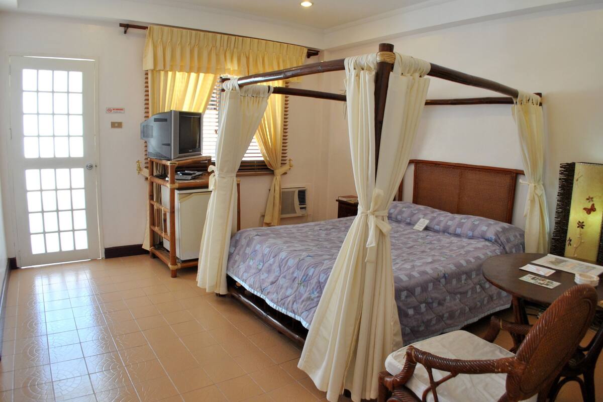 A bedroom with a four-poster bed, a television on a stand, a chair, and a small table.