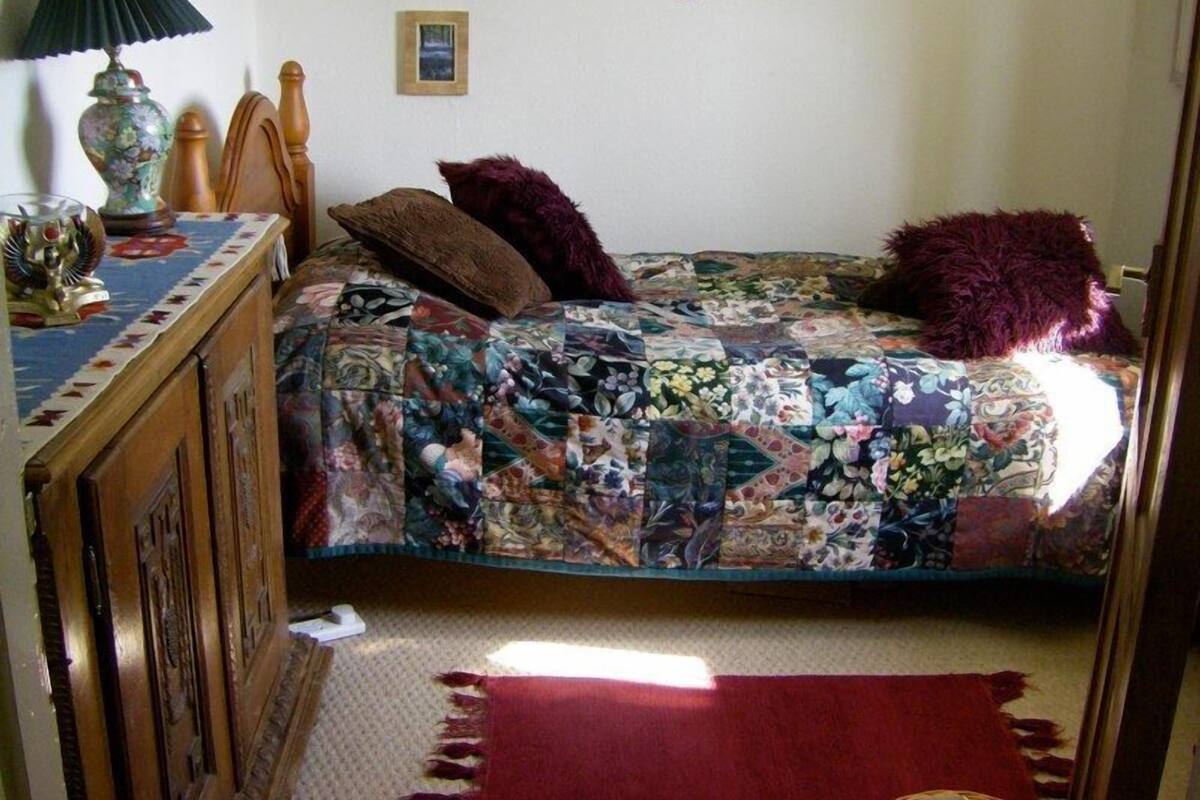 A bedroom with a bed covered in a floral quilt, a wooden dresser with a lamp, and a small framed picture on the wall.