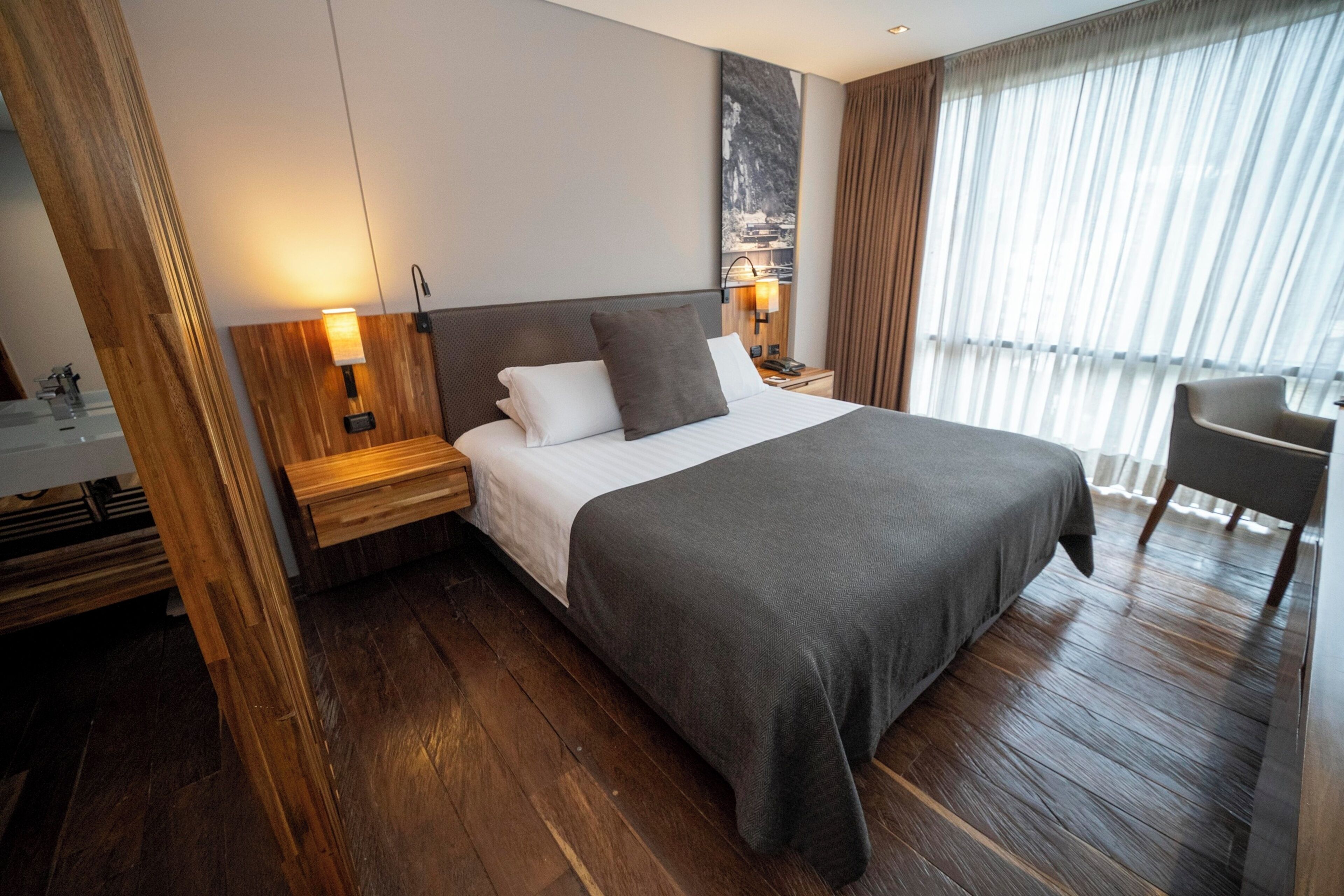 A hotel room with a large bed, wooden headboard, a nightstand, a bedside lamp, a chair, and a window with curtains.