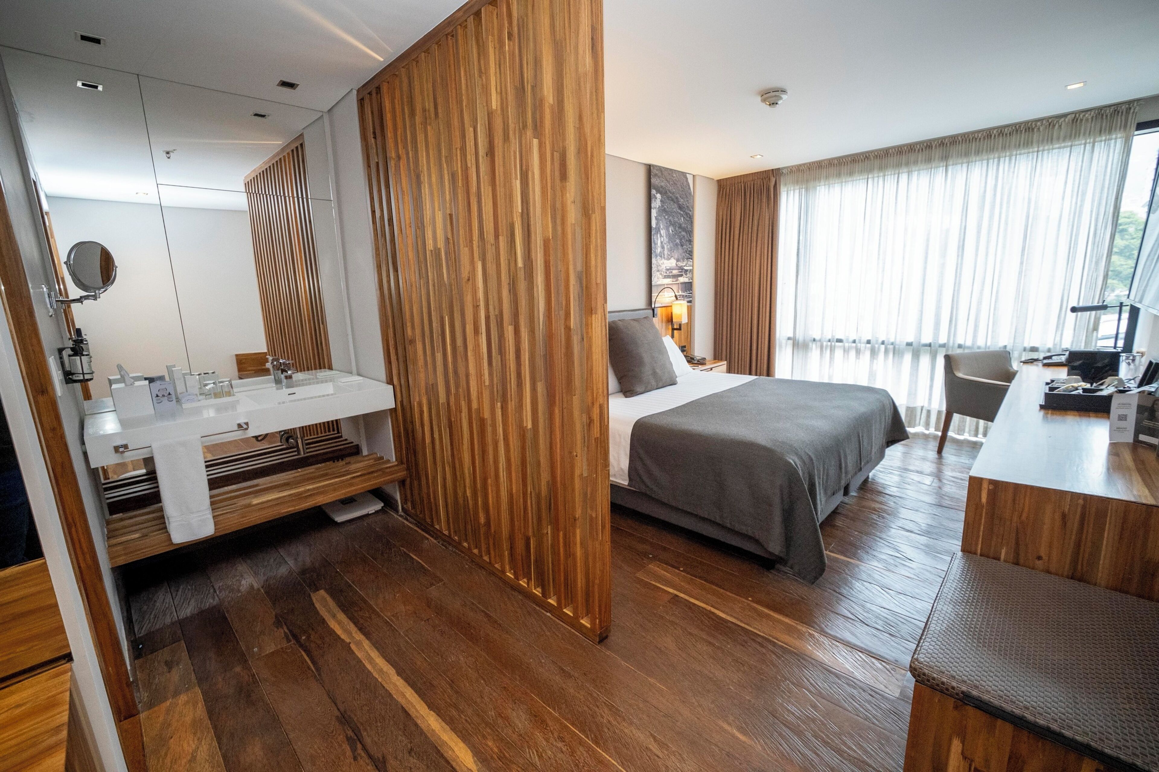 A modern hotel room with a wooden floor, a bed with grey bedding, a desk with a chair, a bathroom with a mirror and sink, and a large window with curtains.