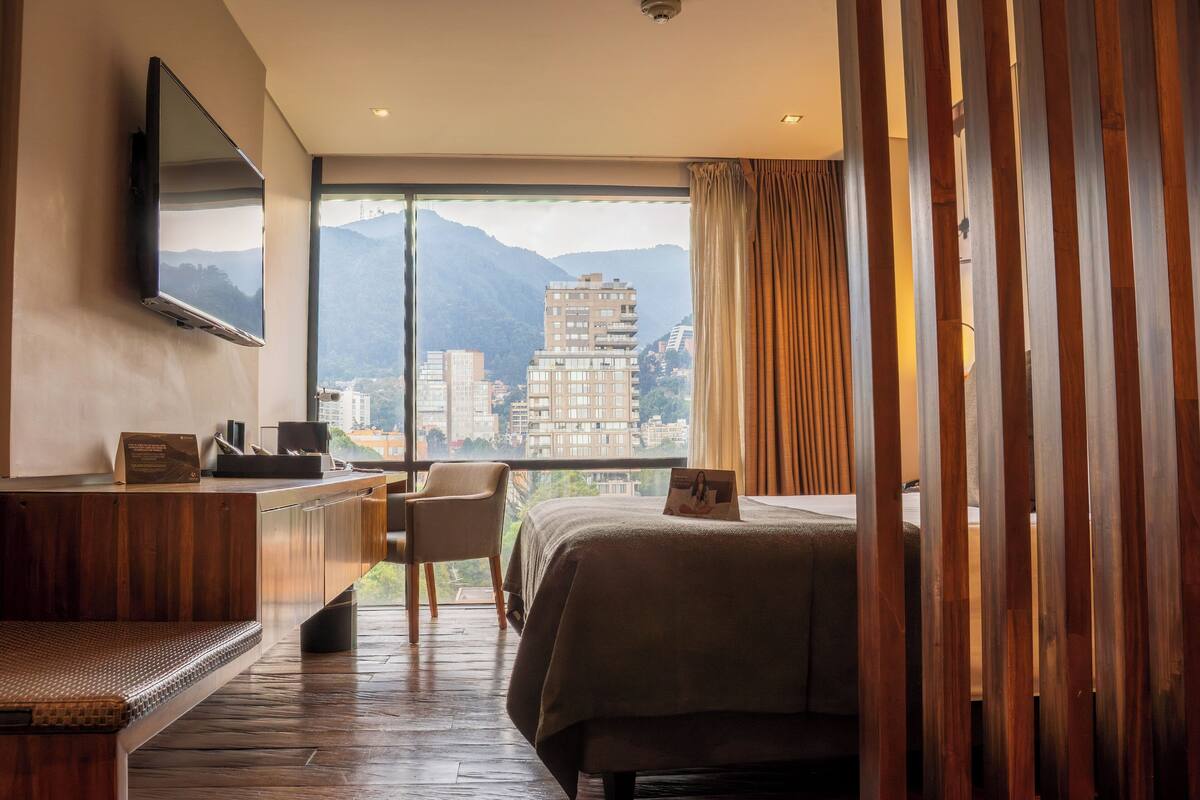 Une chambre d’hôtel équipée d’un lit, d’un bureau, d’une chaise, d’une télévision et d’une grande fenêtre donnant sur les montagnes.