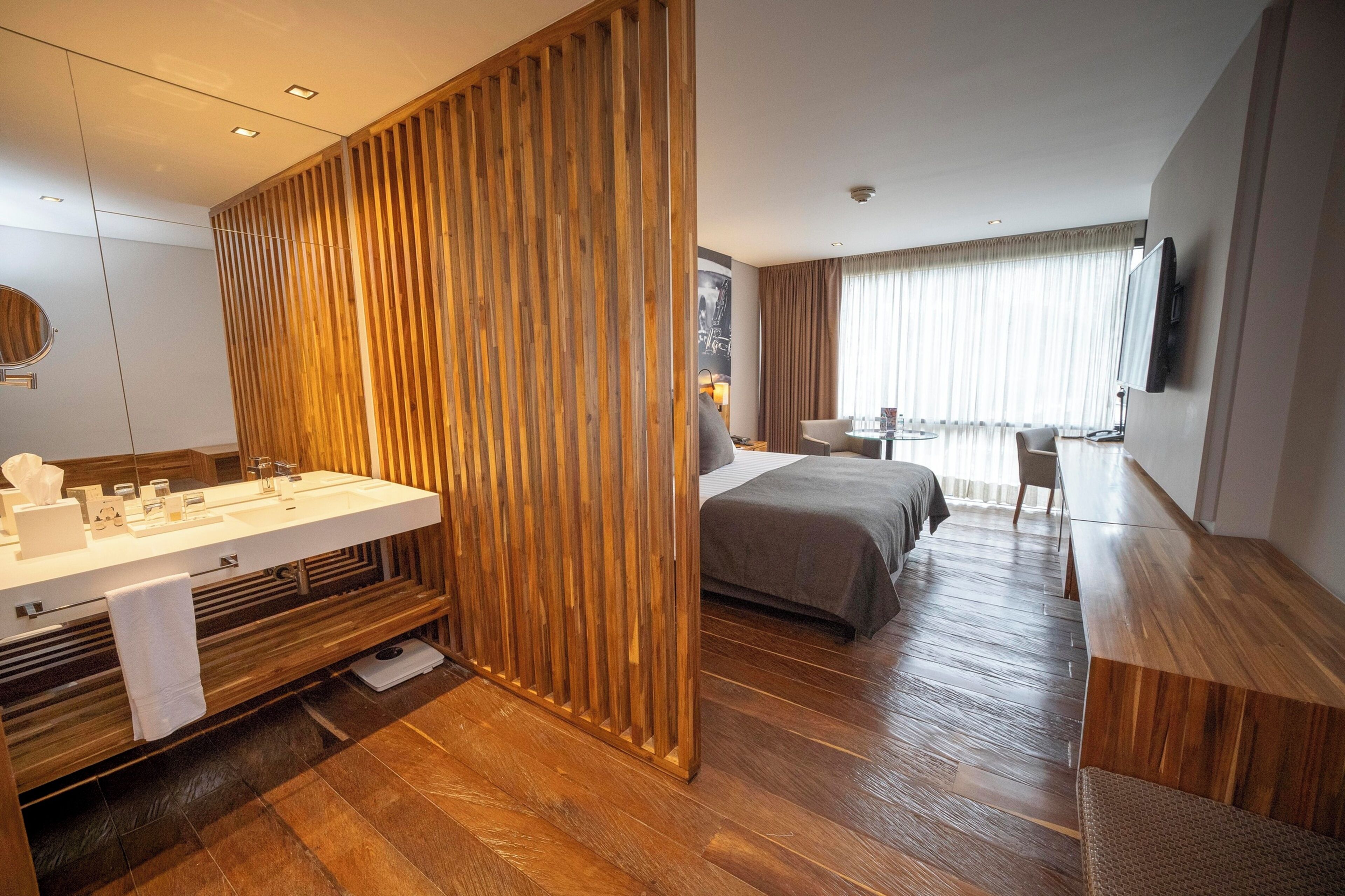 A hotel room with a wooden floor, a bed, a desk with a chair, a bathroom with a sink, and a large window with curtains.