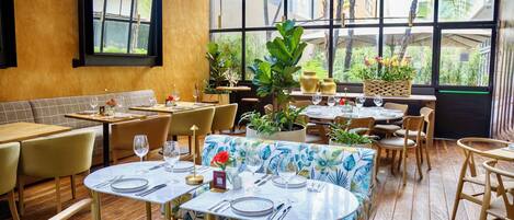 Interior de un restaurante con mesas y sillas de madera, grandes ventanales con vistas a zonas verdes y plantas en macetas.