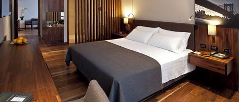 A hotel room with a large bed, wooden flooring, a desk with a telephone, and a nightstand with a lamp.