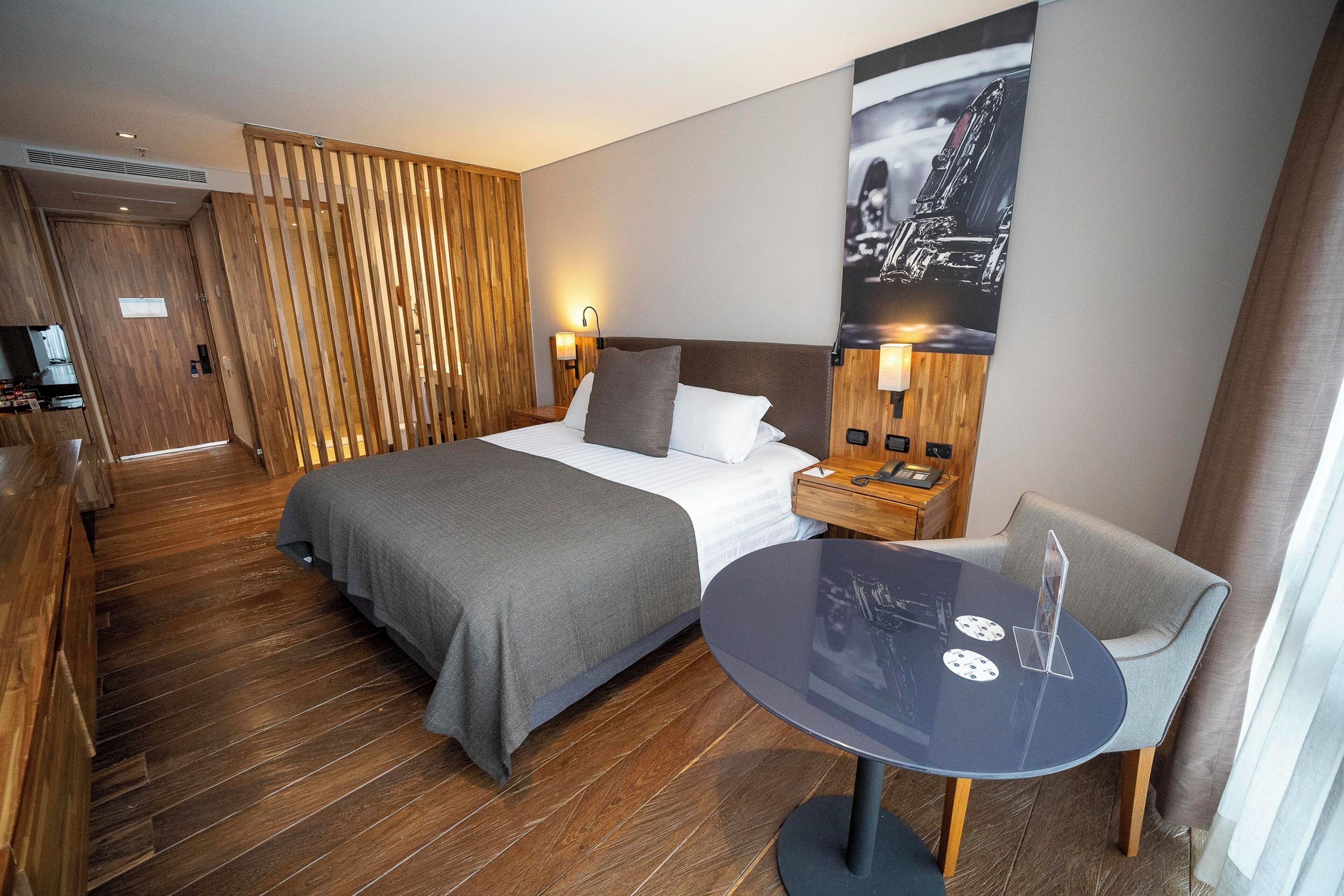 Quarto de hotel com cama, mesa de cabeceira, cadeira e uma pequena mesa com tampo de vidro.