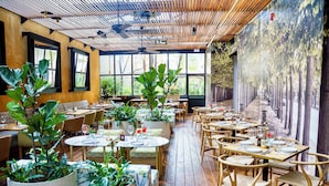 Ein Restaurant mit Holzböden, runden Tischen und Stühlen. Große Kübelpflanzen und ein Wandgemälde sind ebenfalls vorhanden.