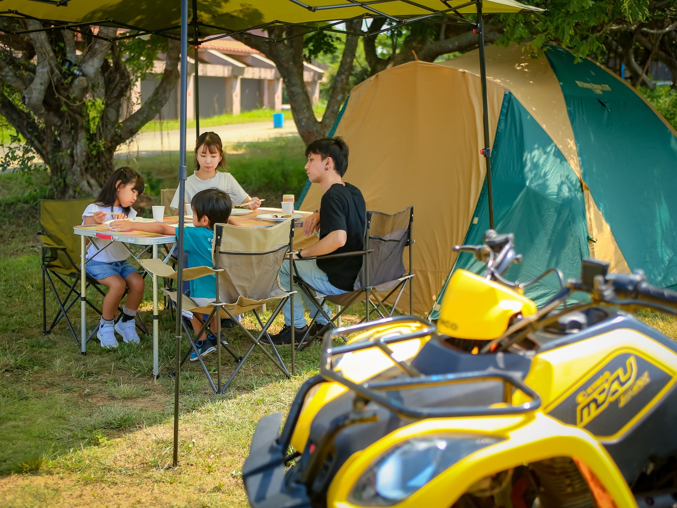 Een gezin dineert aan een buitentafel onder een luifel, met een tent en een gele quad in de voorgrond.