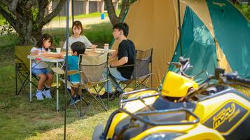 Een gezin dineert aan een buitentafel onder een luifel, met een tent en een gele quad in de voorgrond.